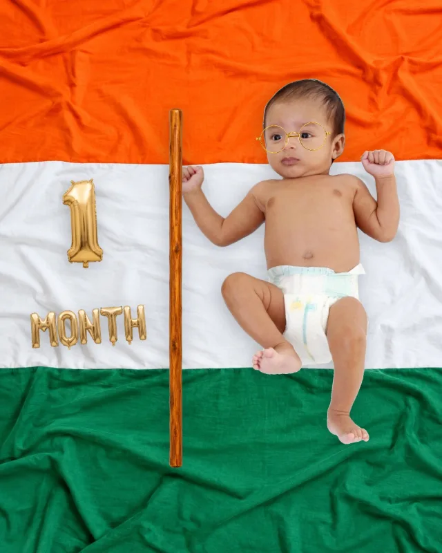 Baby in round Gandhi glasses lying across the Indian tricolor flag holding a small flagpole, beside a gold milestone sticker