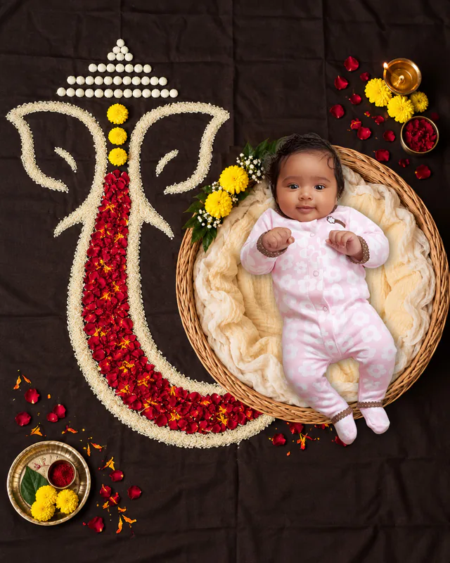 Baby in a woven basket beside a Ganesh motif made from rice, rose petals and marigolds