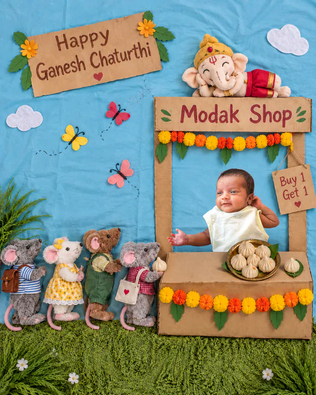 Baby at a cardboard modak stall with plush mouse customers and a Ganesha toy watching over the shop