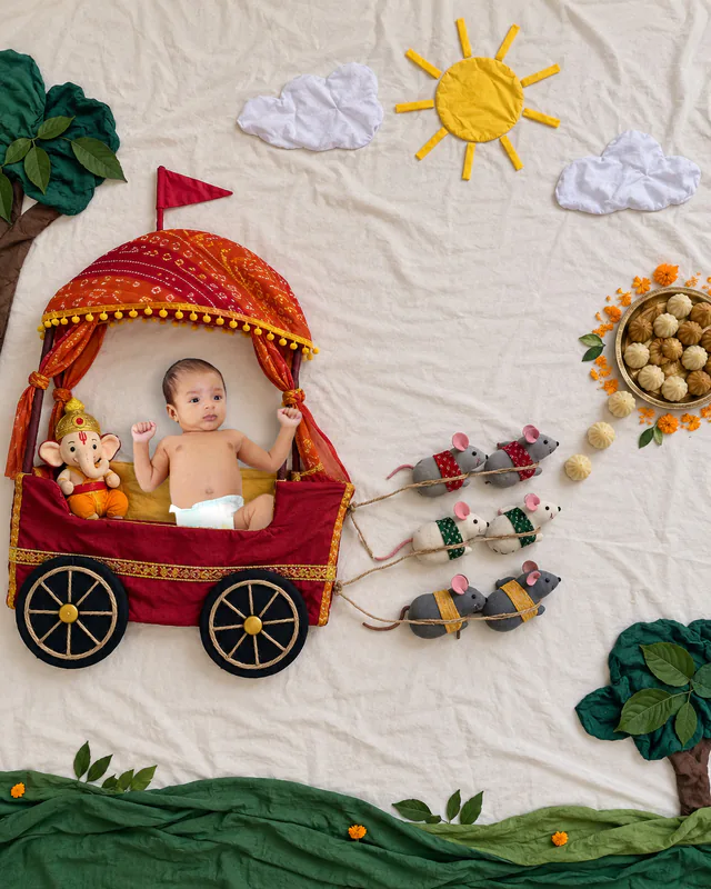 Baby riding in a festive cloth rath pulled by toy mice beside a plush Ganesha and modaks