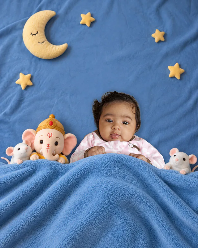 Baby peeking from a blue blanket beside a plush Ganesha and mouse toys under a starry sky