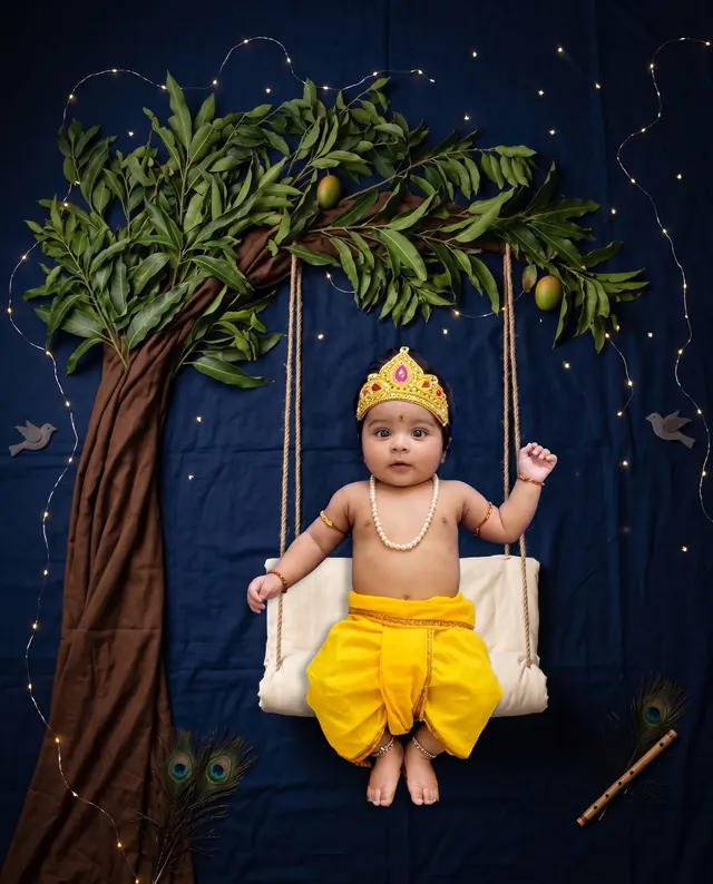 Baby in a Krishna outfit on a white swing framed by leafy branches against a night sky