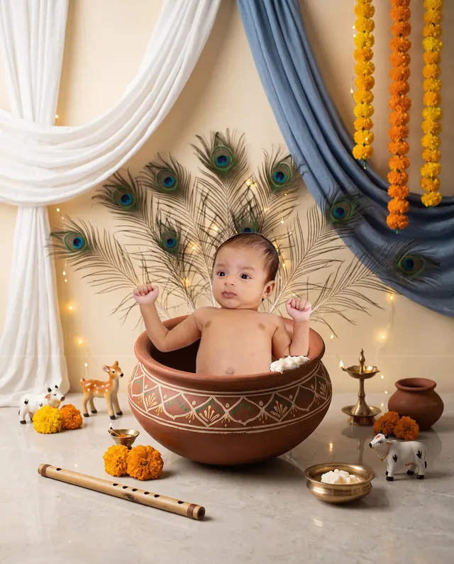 Baby peeking out of an earthen matka with peacock feathers, cream drapes and blue fabric