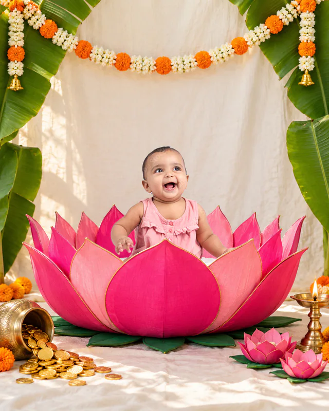 Smiling baby sitting inside a pink lotus beneath marigold garlands and banana leaves with gold coins beside the flower